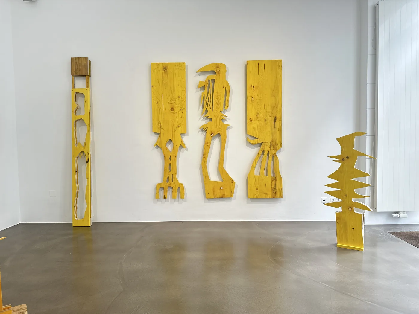 Wide installation view of five formwork-board sculptures on a gallery wall and floor, Cosmic Trigger – Respawn, Bildraum 07, Ernst Koslitsch