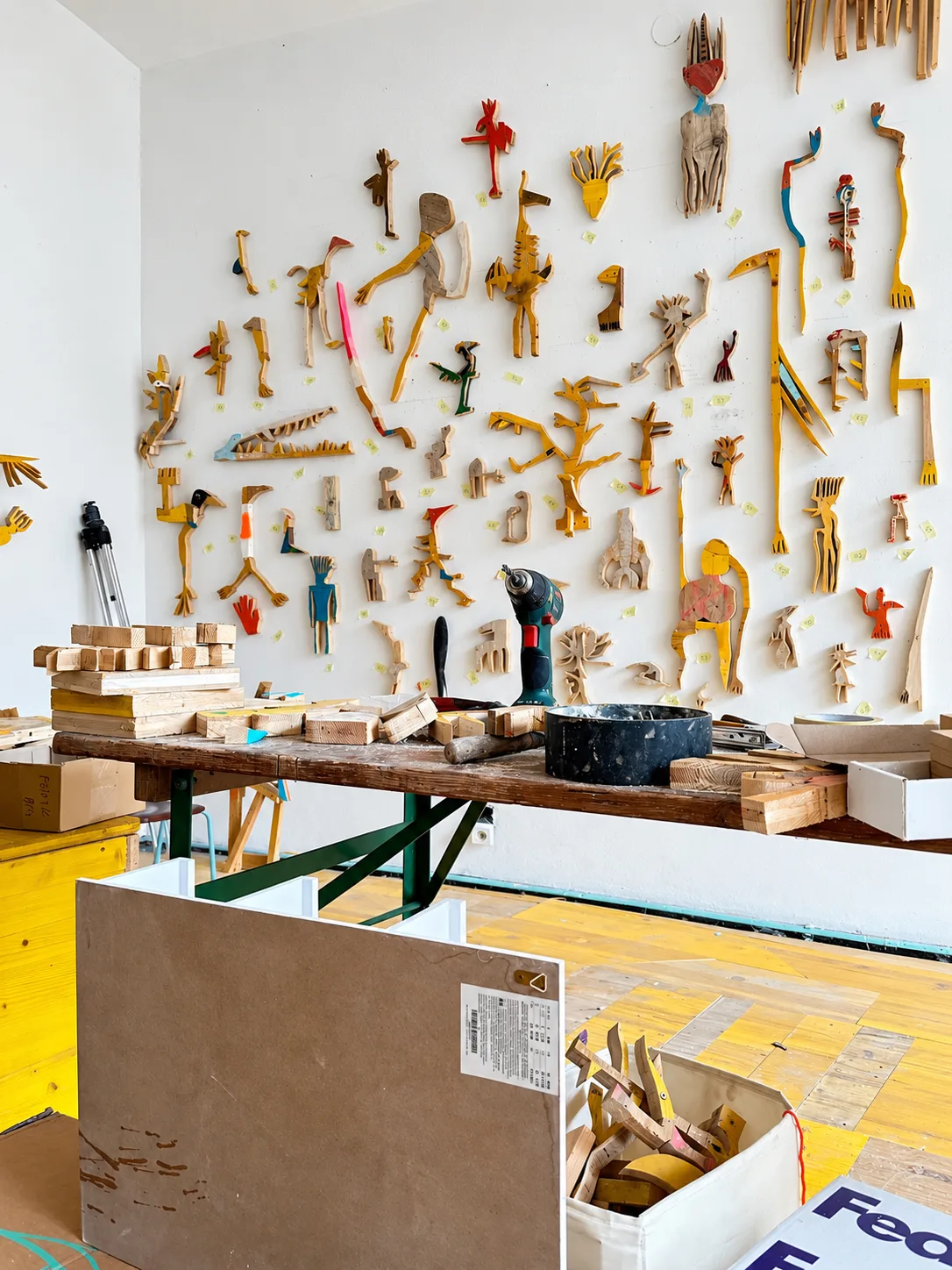 Workbench with tools, wood offcuts and packing crates below a wall of small painted wood figures, studio view, Ernst Koslitsch