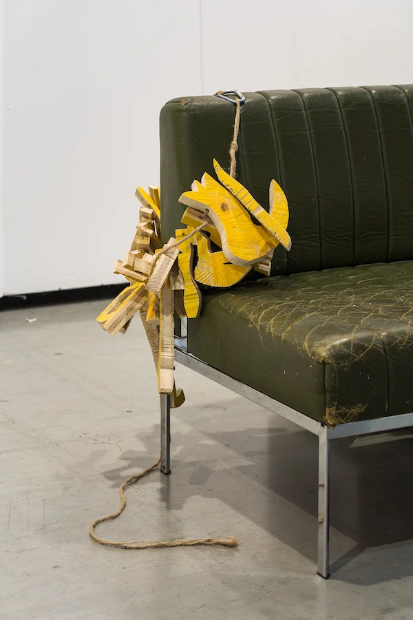 Yellow wood sculpture hanging on a sofa arm, Spark Art Fair booth, Ernst Koslitsch